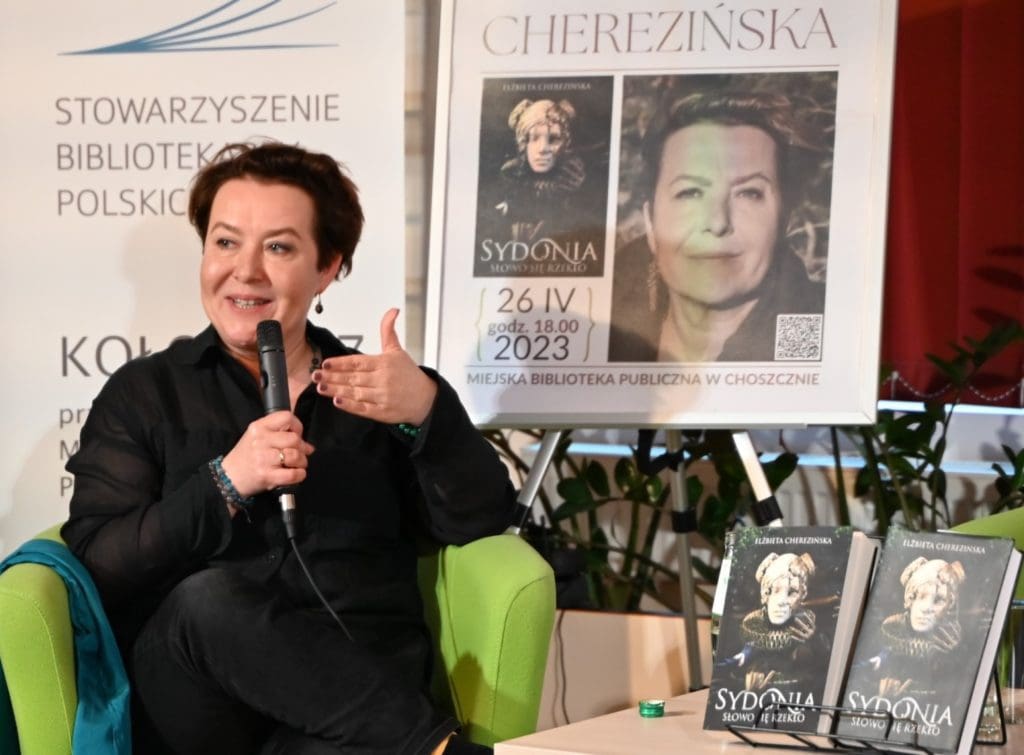 Elżbieta Cherezińska, pisarka siedzi w fotelu na tle baneru i plakatu, w prawej ręce trzyma mikrofon. Na stoliku obok stoją książki.