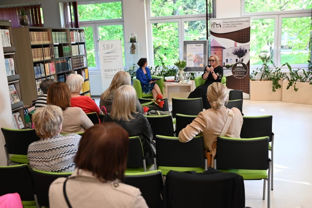 W czytelni biblioteki siedzi grupa osób na spotkaniu autorskim. W głębi siedzi pisarka Iwona Małgorzata Żytkowiak z prowadzącą spotkanie bibliotekarką Wiolettą Witkowską.