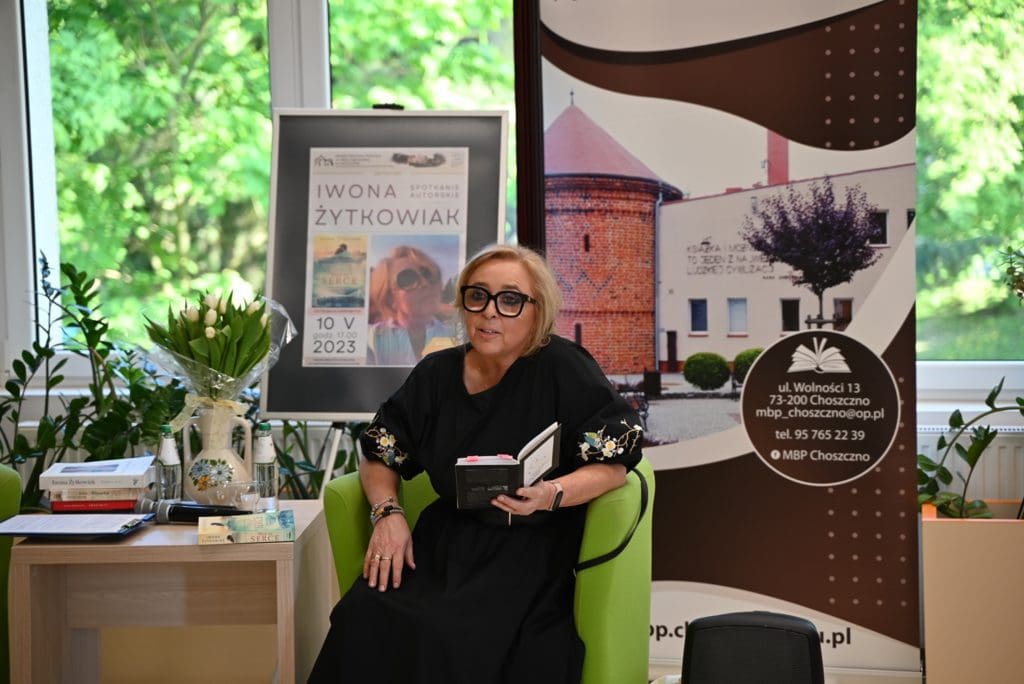 Na tle okien,  Iwona Małgorzata Żytkowiak , ubrana w czarną sukienkę, siedzi w fotelu z książką w ręku. Obok na stoliku stoją w wazonie tulipany, leżą książki i mikrofon. W tle stoi plakat i baner biblioteki.
