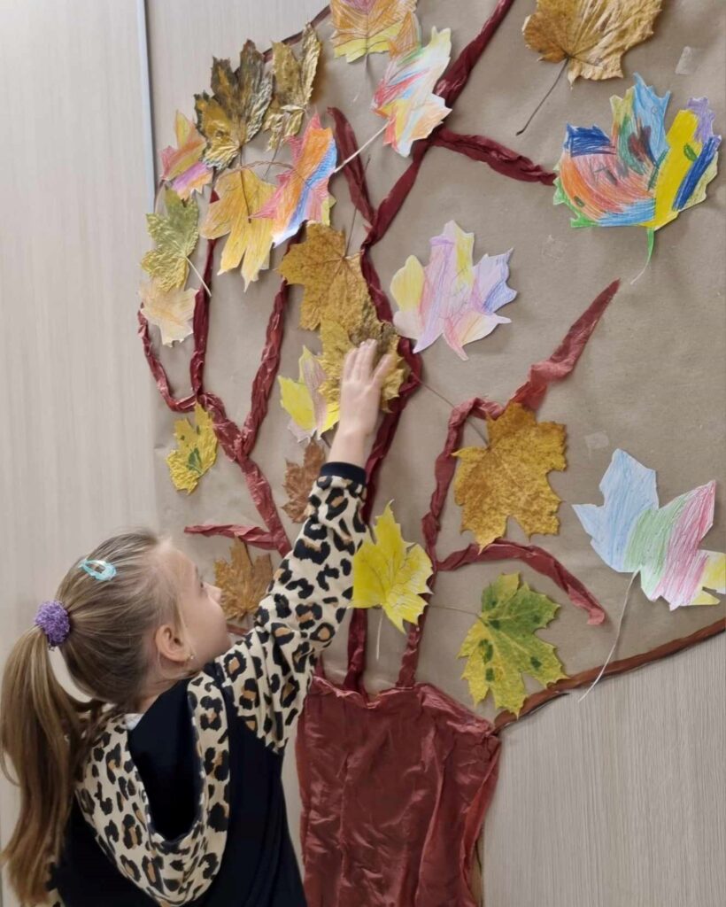 Dziewczynka przyczepia liść do namalowanego drzewa.