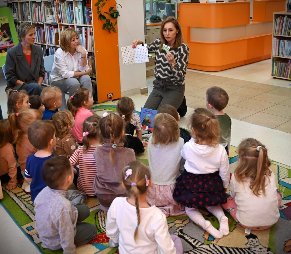 Bibliotekarka Izabela Szcyż pokazuje dzieciom siedzącym na dywanie  kartkę z naklejkami. Obok na niskich krzesełkach siedzą przedszkolanki.