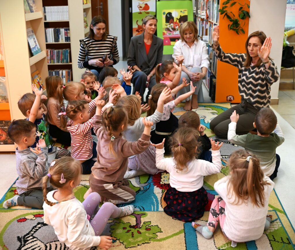 Bibliotekarka Izabela Szcyż pokazuje dzieciom gest : uniesione ręce, zgięte w łokciach, widoczne wnętrze dłoni.  Część dzieci siedzących na dywanie ją naśladuje. Obok na niskich krzesełkach siedzą przedszkolanki.