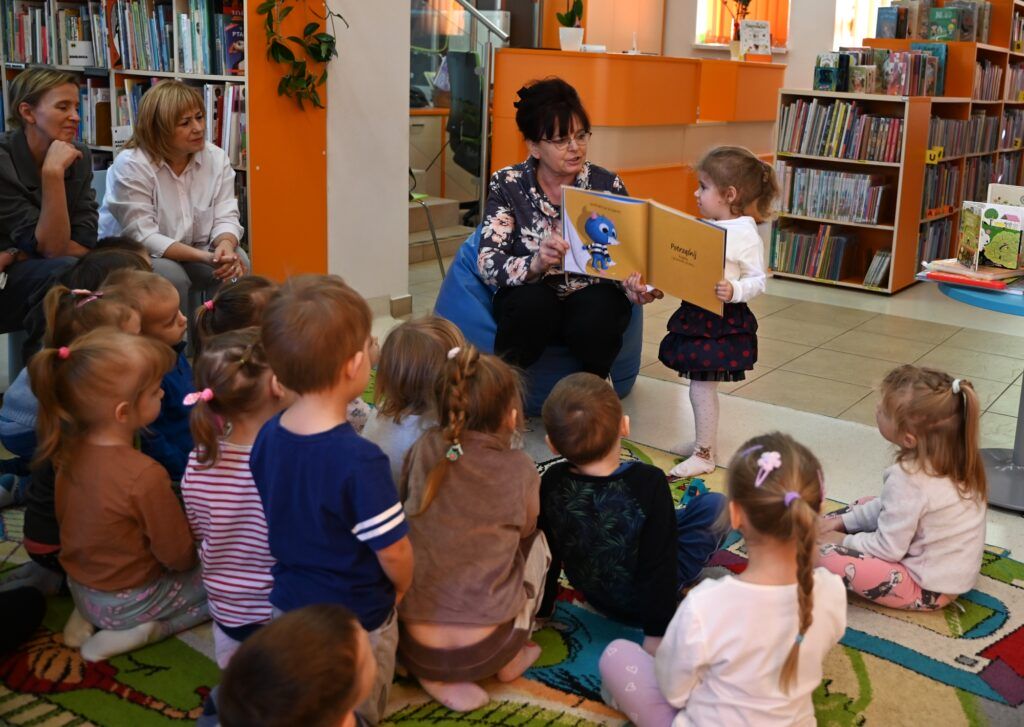 Bibliotekarka Irena Kozłowska pokazuje dziewczynce z bliska obrazki w książce. Reszta dzieci siedzi na dywanie. Obok na niskich krzesełkach siedzą przedszkolanki.