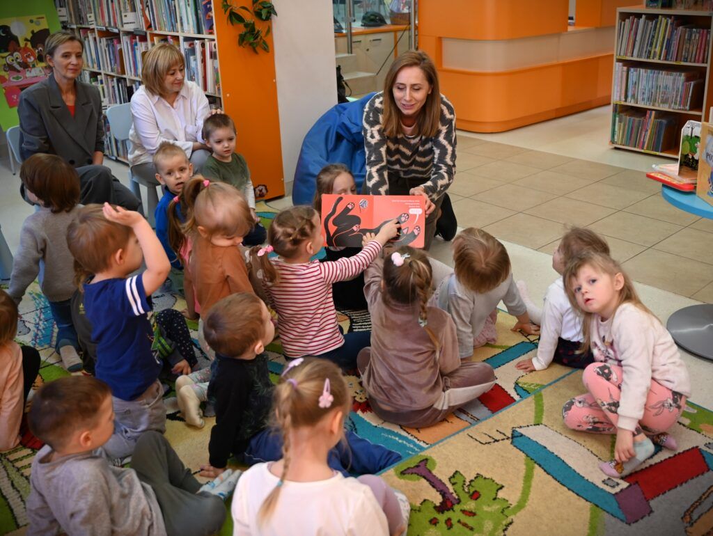 Bibliotekarka Izabela Szcyż pokazuje dzieciom siedzącym na dywanie książkę.   Obok na niskich krzesełkach siedzą przedszkolanki.
