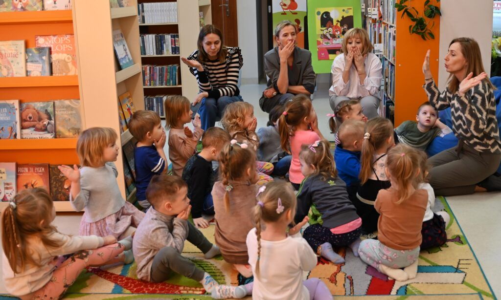 Bibliotekarka Izabela Szcyż pokazuje dzieciom "puszczanie buziaków".  Część dzieci siedzących na dywanie ją naśladuje. Obok na niskich krzesełkach siedzą przedszkolanki, które też naśladują ten gest.
