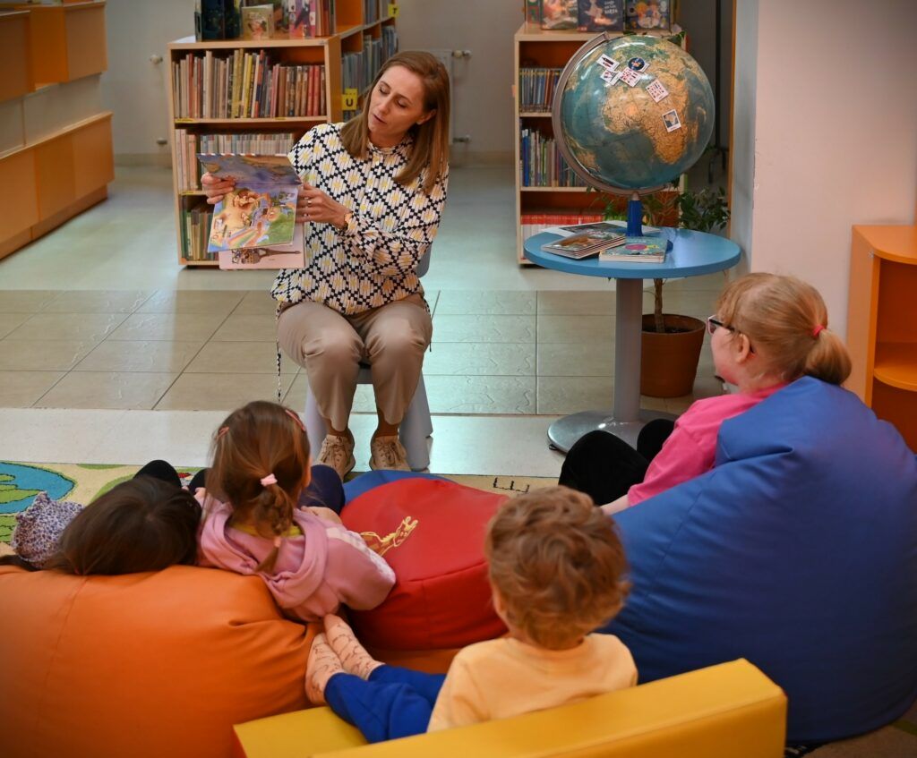 Bibliotekarka pokazuje dzieciom książkę. Obok niej na stoliku stoi globus.