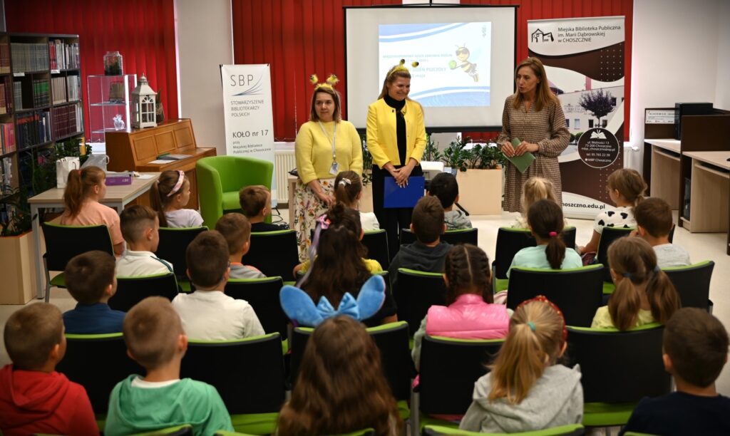 Od lewej stoją: Agnieszka Żygadło,  Monika Pilch-Cieślak  i bibliotekarka Izabela Szczyż przed siedzącymi dziećmi. Prelegentki mają na głowach przepaski z pszczelimi czułkami.