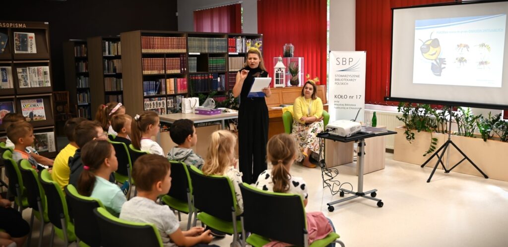 Jedna z prelegentek stoi z kartką w ręku przed grupą dzieci, druga siedzi z tyłu. Obie mają na głowach przepaski z pszczelimi czułkami. Obok stoi rzutnik i ekran, na którym wyświetlony jest obrazek pszczoły.