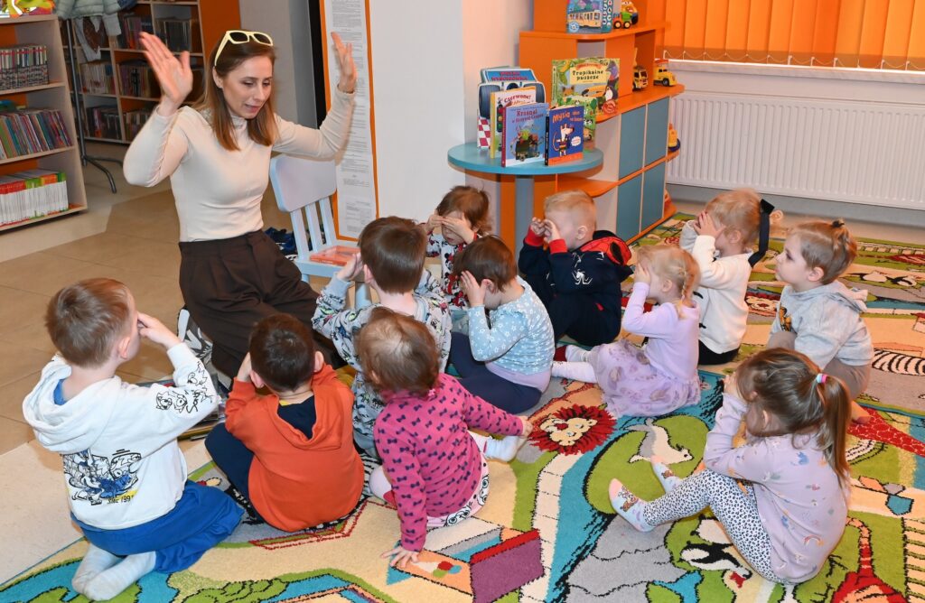 Bibliotekarka Izabela Szczyż klęczy przed dziećmi z podniesionymi rękami. Dzieci zakrywają oczy dłońmi.