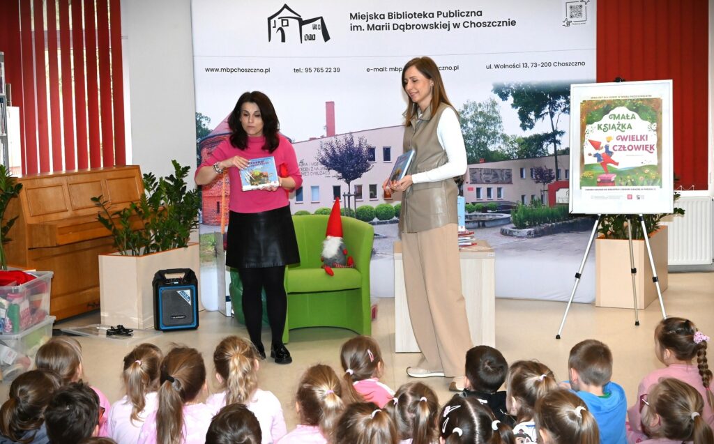  Lucyna Brzezińska-Eluszkiewicz i bibliotekarka Izabela Szczyż stoją z książkami w rękach. Z prawej strony plakat akcji "Mała książka - wielki człowiek". Z przodu widać głowy dzieci.