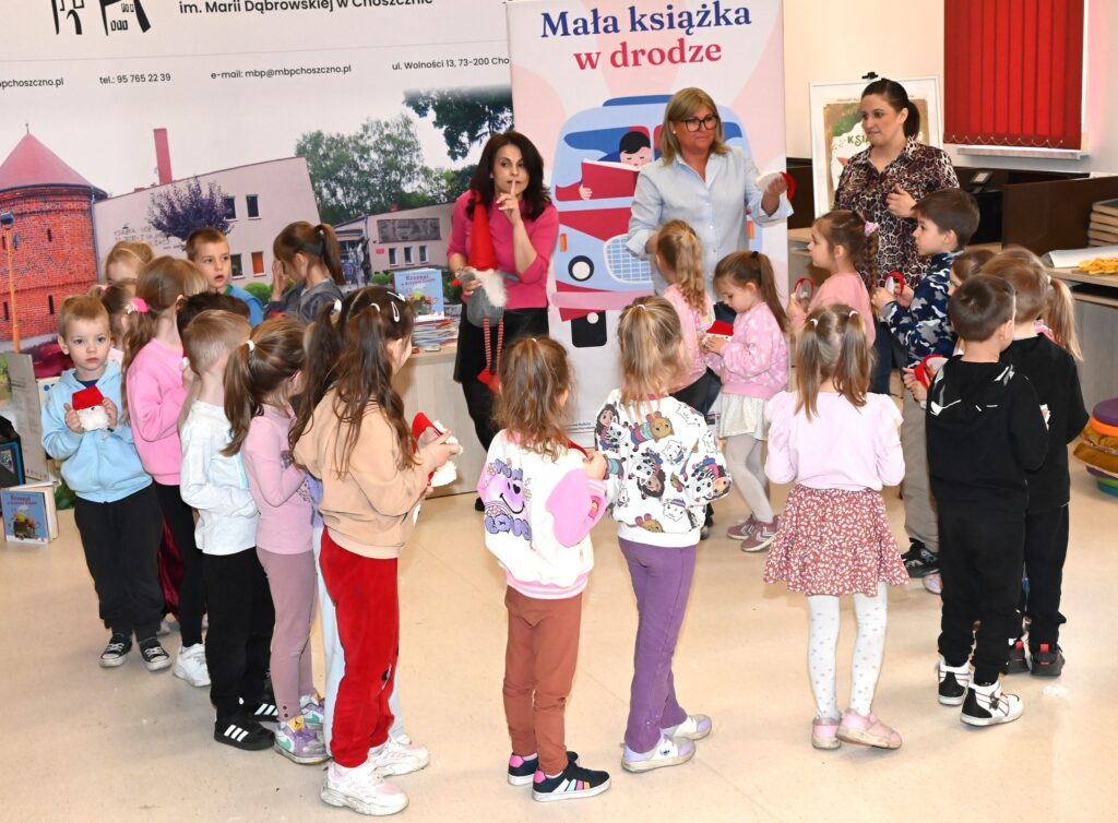 Dzieci, przedszkolanki i Lucyna Brzezińska-Eluszkiewicz stoją w kręgu. Lucyna trzyma przy ustach palec.