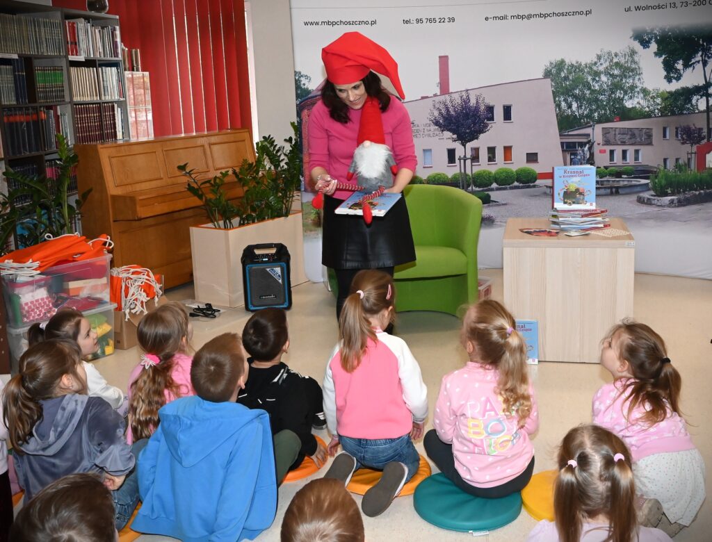  Lucyna Brzezińska-Eluszkiewicz siedzi w fotelu przed dziećmi siedzącymi na poduszkach. N a głowie ma czerwoną krasnoludkową czapkę. W ręku trzyma krasnoludka i książkę, z której czyta.