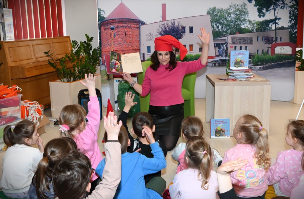  Lucyna Brzezińska-Eluszkiewicz siedzi w fotelu przed dziećmi siedzącymi na poduszkach. N a głowie ma czerwoną krasnoludkową czapkę. W ręku trzyma książkę, z którą pokazuje dzieciom