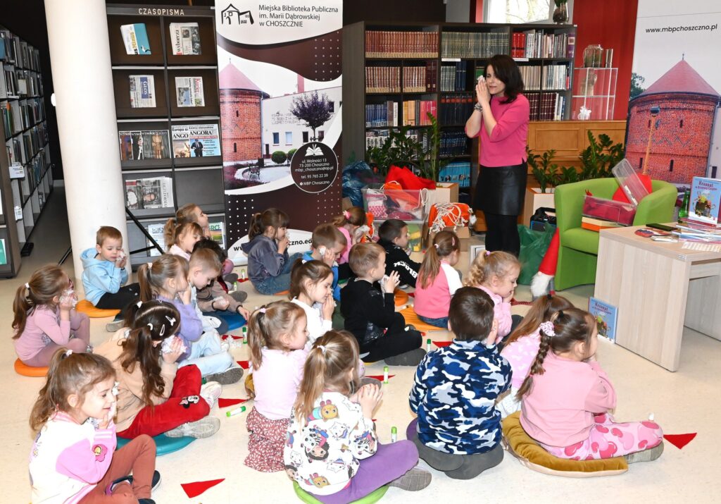  Lucyna Brzezińska-Eluszkiewicz stoi przed dziećmi siedzącymi na poduszkach. Trzyma złożone ręce przy twarzy.