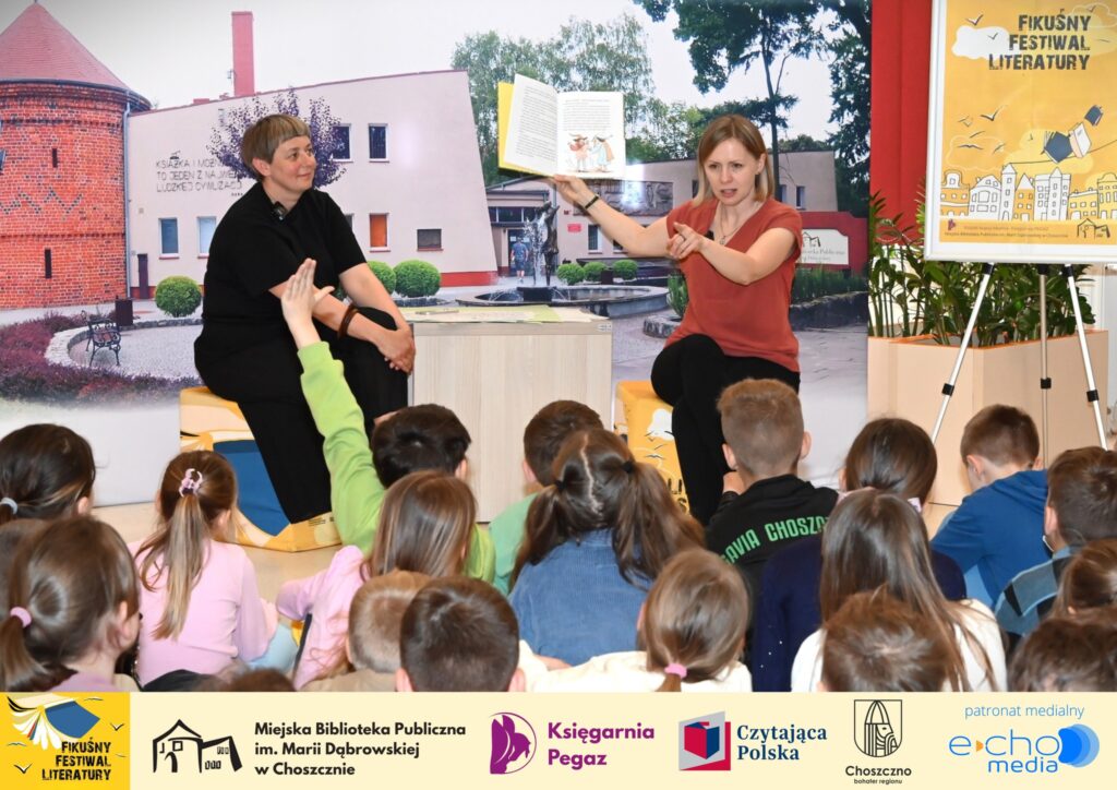 Przed widownią dzieci siedzą od lewej Małgorzata Narożna i Magdalena Kiełbowicz, która w górze trzyma otwarta książkę.