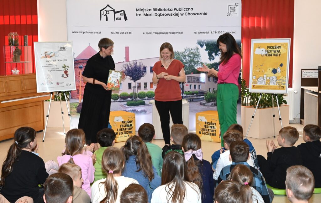 Przed publicznością dzieci stoją od lewej: MAłgorzata Narożna z książką w ręku, Magdalena Kiełbowicz i Katarzyna Jezierska, dyrektorka biblioteki.