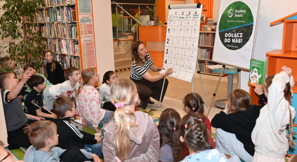 Bibliotekarka Izabela Szczyż kuca przy tablicy z punktacją. Podlicza punkty. Przed nią dzieci.