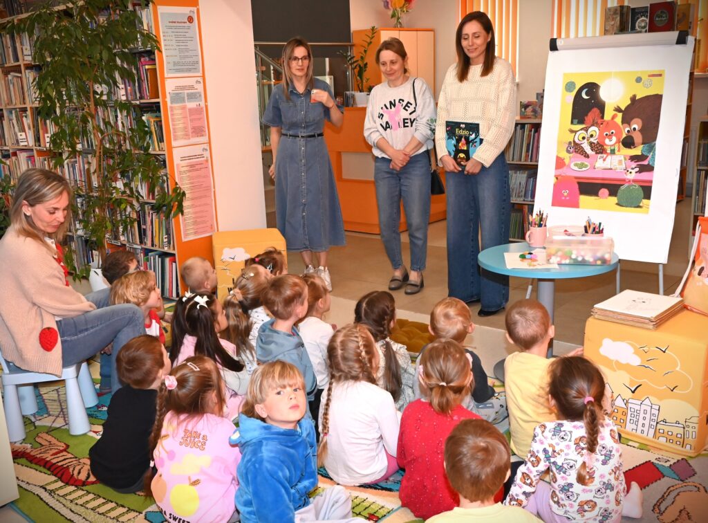 Bibliotekarka Iza Szczyz oraz przedszkolanki stoją przed siedzącymi na dywanie dziećmi