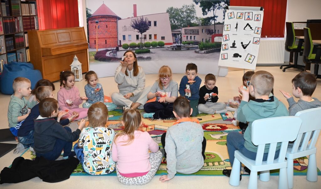 Dzieci siedzą w kręgu z bibliotekarką Izą Szczyż i dmuchają na piórka.