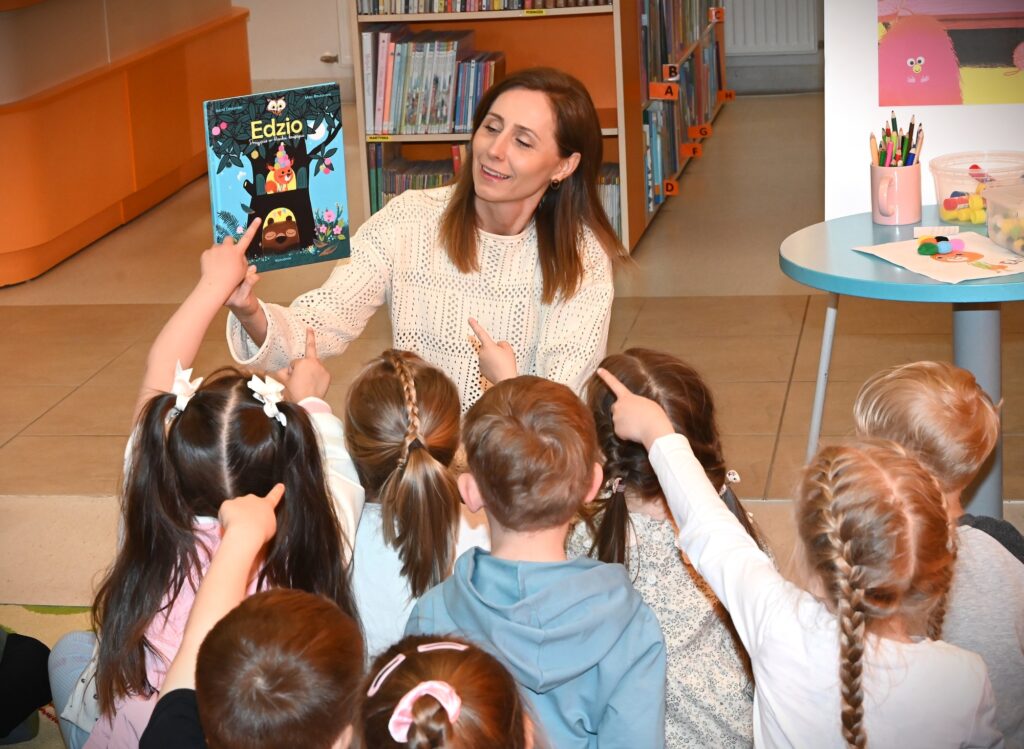 Bibliotekarka Iza Szczyz pokazuje dzieciom książkę o Edziu. Dzieci wyciągają ręce.