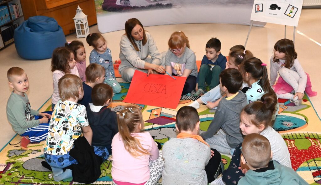 Dzieci siedzą w kręgu z bibliotekarką Izą Szczyż, która trzyma w ręku dużą czerwoną kartkę z napisem "cisza".