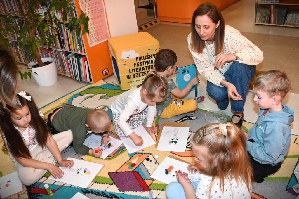 Bibliotekarka Iza Szczyz kuca przy dzieciach, które przyklejają małe pomponiki do rysunków.