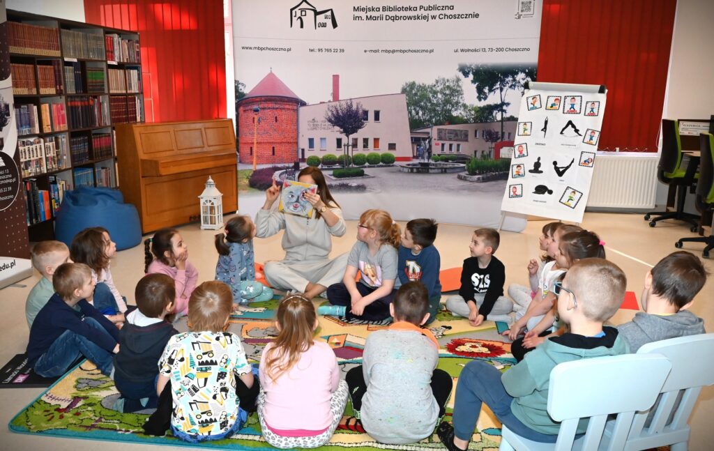 Dzieci siedzą w kręgu z bibliotekarką Izą Szczyż, która trzyma przed twarzą książkę.
