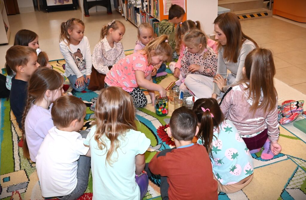 Dzieci i bibliotekarka Izabela Szczyz siedzą w kole na dywanie. Jedna z dziewczynek wyciąga ze słoika pełnego kolorowych małych pomponów jeden z nich.