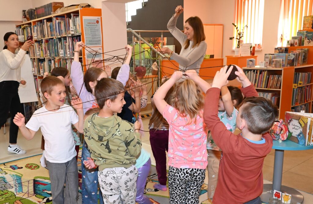 Dzieci i bibliotekarka Izabela Szczyż stoją w kole. Przeplatają miedzy sobą nić.