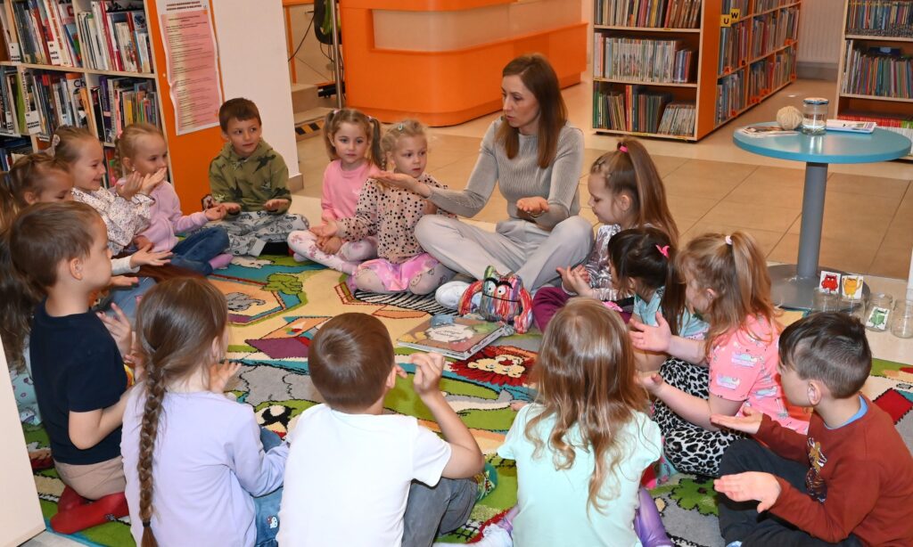 Dzieci i bibliotekarka Izabela Szczyz siedzą w kole. Wzyscy trzymają ręce dłońmi do góry.