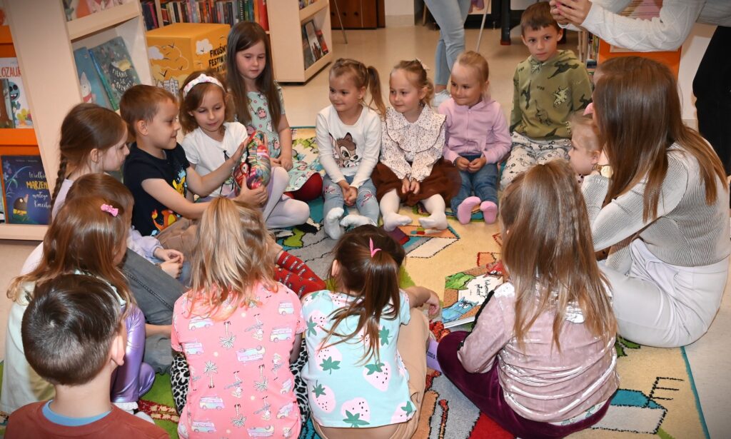 Dzieci i bibliotekarka Izabela Szczyz siedzą w kole. jeden z chłopców trzyma w ręku maskotkę wielokolorowego potwora.