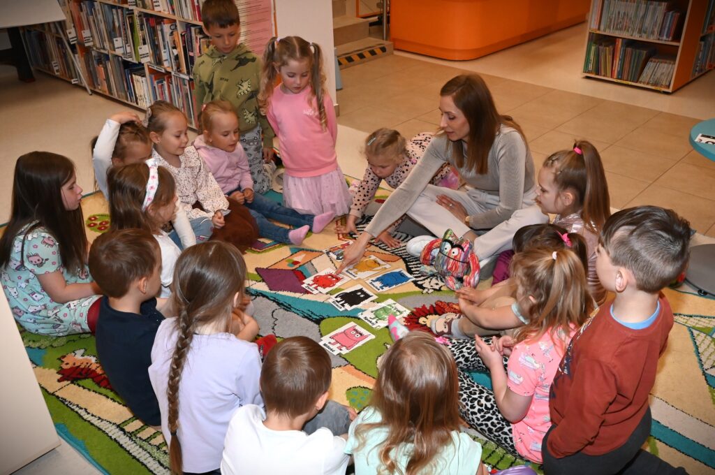 Dzieci i bibliotekarka Izabela Szczyz siedzą w kole. po środku leżą rysunki z potworami w różnych kolorach. bibliotekarka wskazuje czerwonego. obok niej jest wielokolorowa maskotka potwora.