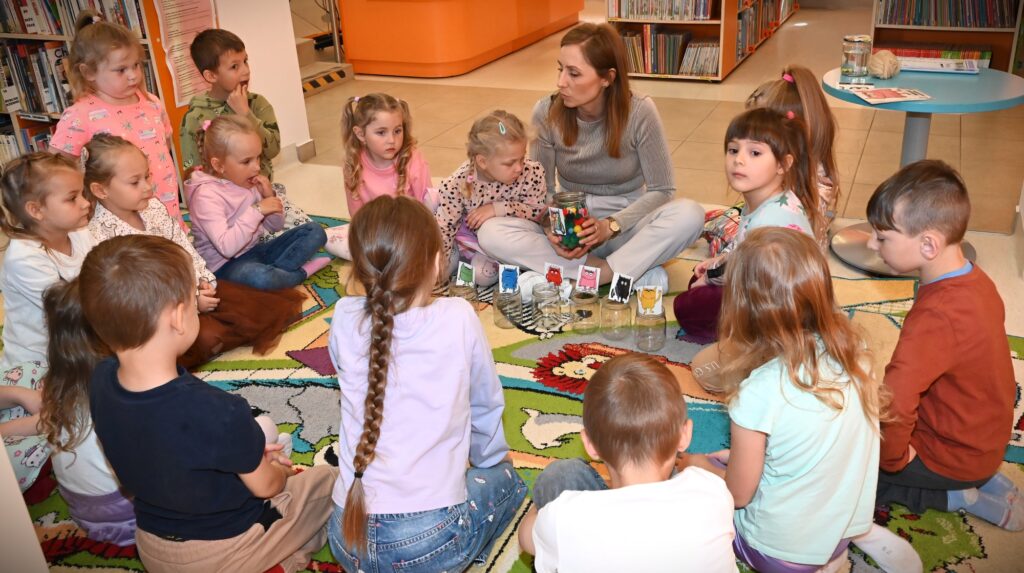 Dzieci i bibliotekarka Izabela Szczyz siedzą w kole.  Bibliotekarka trzyma w rekach słoik pełen kolorowych pomponików. Przed nią stoją słoiki oznaczone rysunkami potworów w różnych kolorach.