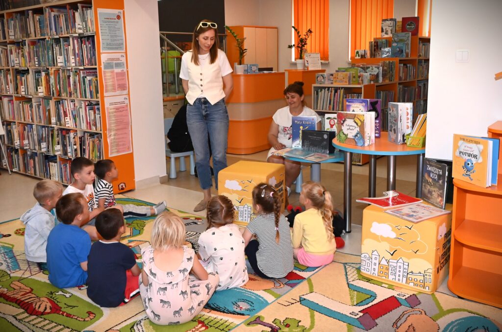 Bibliotekarka Izabela Szczyż stoi przed dziećmi siedzącymi na dywanie. Obok niej stolik z książkami, przy którym siedzi przedszkolanka.