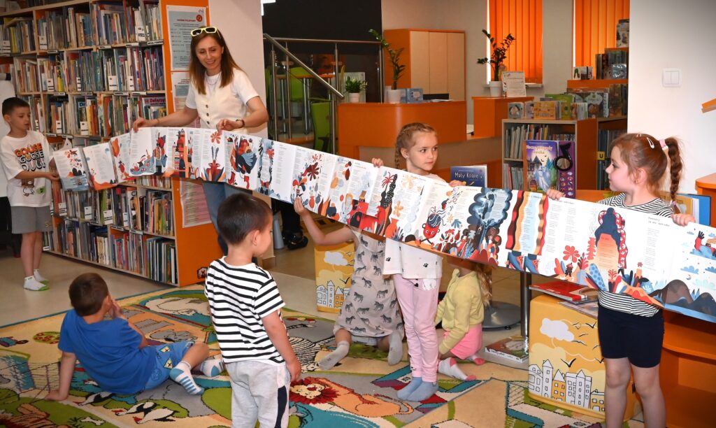 Bibliotekarka Izabela Szczyż razem z dziećmi stoją trzymając rozłożoną wzdłuż książkę harmonijkę.