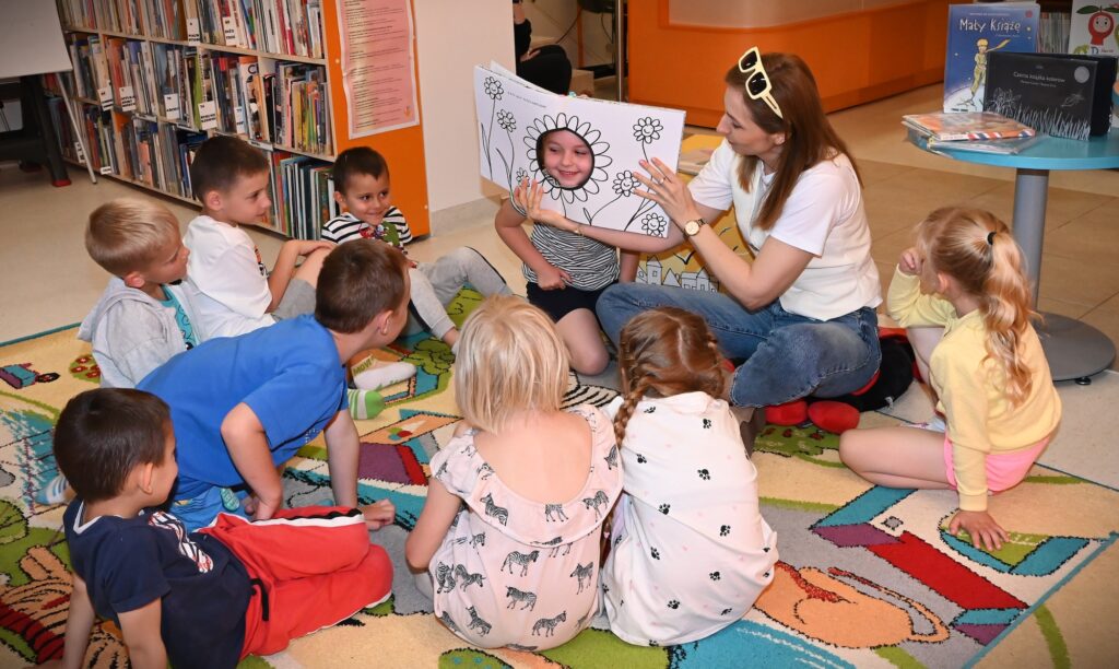 Bibliotekarka Izabela Szczyż trzyma otwartą książkę, w której jest otwór z obrysem kwiatka. W otworze tkwi twarz dziewczynki. Wkoło siedzą dzieci.