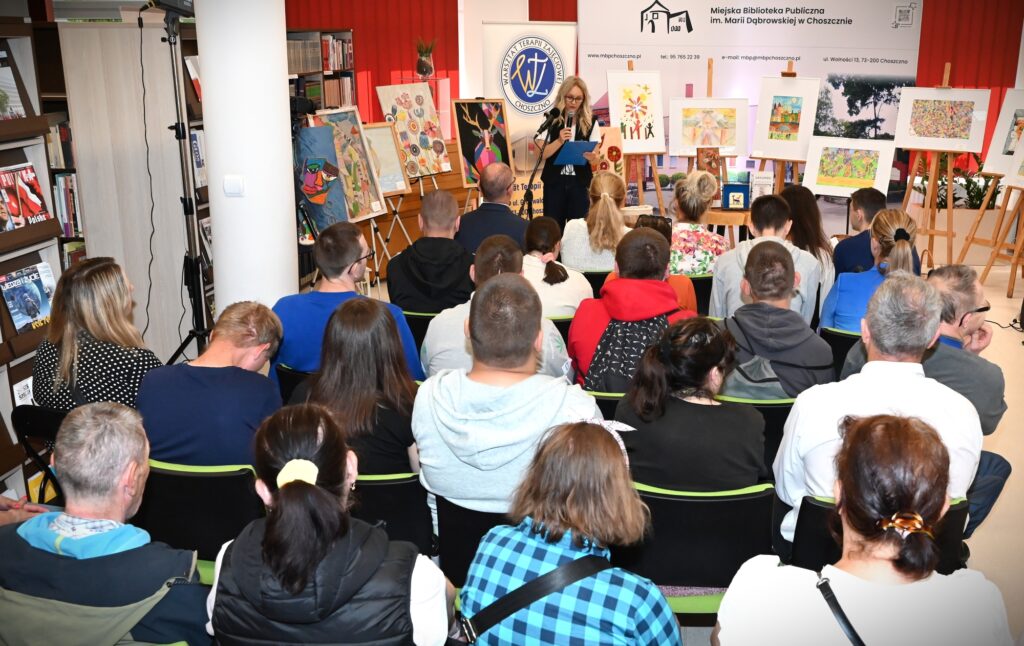 Elwira Najdora stoi przed publicznością.  Za nią stoją obrazy.