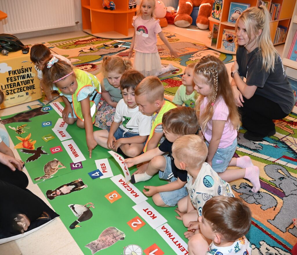 Dzieci siedzą na dywanie. Za nimi siedzi przedszkolanka. Wzdłuż leży zielony karton, na którym są ponumerowane obrazki zwierząt. Dzieci układają ich nazwy. 