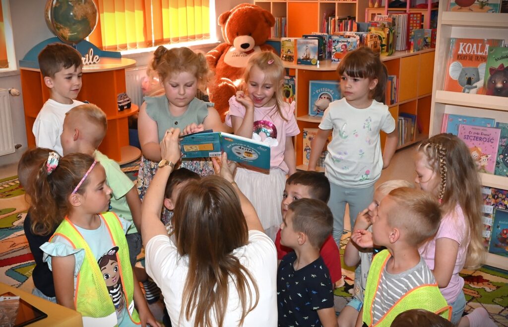 Bibliotekarka Izabela Szczyż pokazuje dzieciom książkę dźwiękową. Jedna z dziewczynek dotyka kartki.