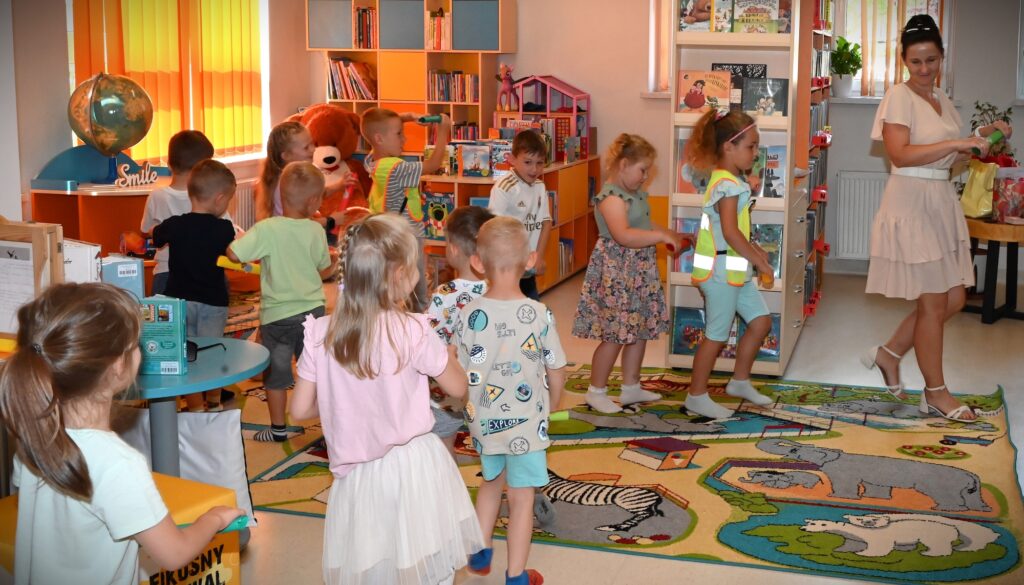 Dzieci idą rządkiem z przedszkolanką. Każdy w ręce trzyma zwinięty karton, który udaje kierownicę roweru.