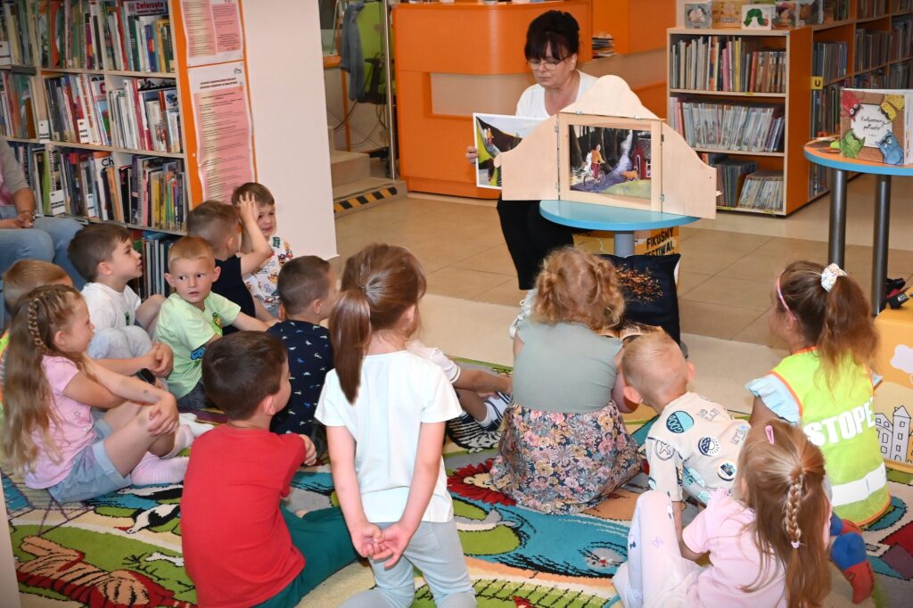Bibliotekarka Irena Kozłowska siedzi przy stoliku. Na stoliku stoi teatrzyk Kamishibai. Bibliotekarka zmienia karty w teatrzyku. Przed nią na dywanie siedzą dzieci.