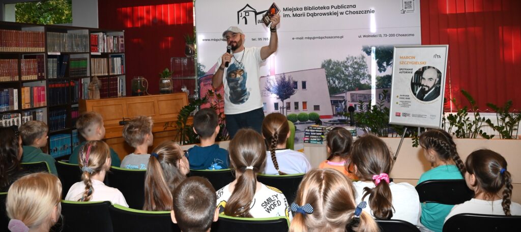 Marcin Szczygielski stoi przed dziećmi, w lewej ręce trzyma swoją książkę.