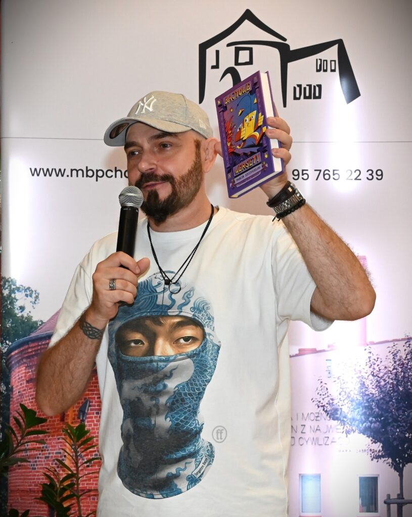 Marcin Szczygielski stoi z mikrofonem i w lewej ręce trzyma swoją książkę.
