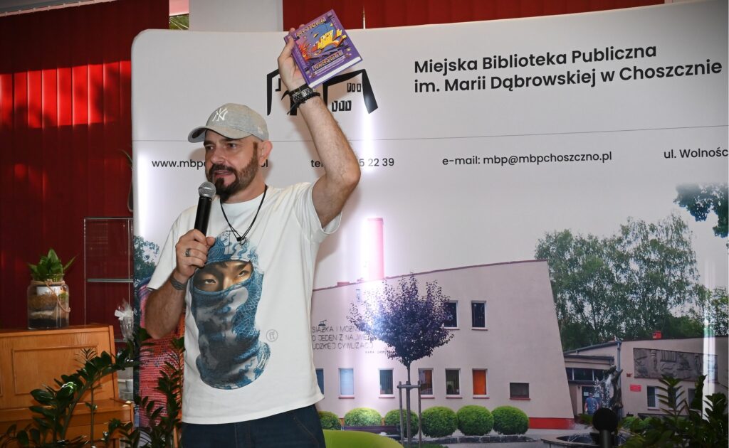 Marcin Szczygielski stoi z mikrofonem i w lewej ręce trzyma swoją książkę.