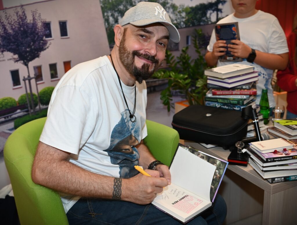 Marcin Szczygielski siedzi w fotelu, Patrzy prosto w kadr. Na kolanach trzyma otwartą książkę, którą podpisuje.