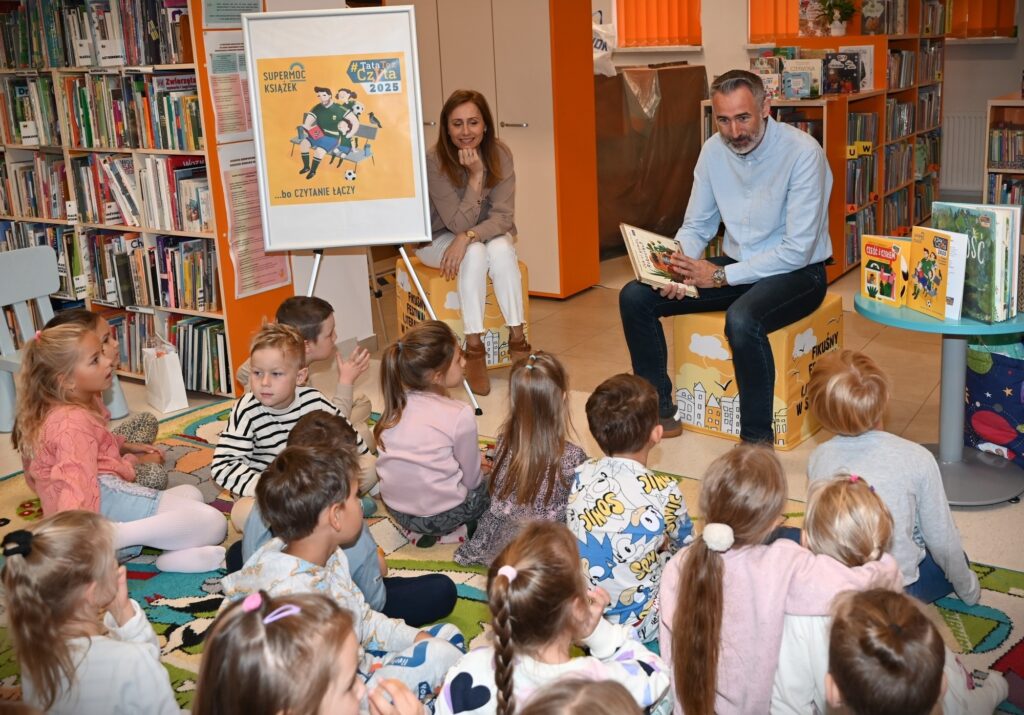 Na pufach siedzą: bibliotekarka Izabela Szczyż oraz pan Piotr z książką w ręku. Przed nimi  siedzą dzieci na dywanie.