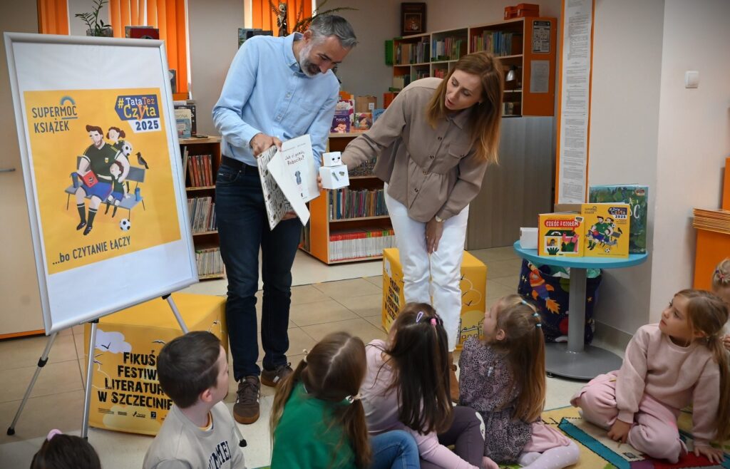 Przed dziećmi stoją pan Piotr i bibliotekarka Izabela Szczy. Mężczyzna trzyma w rękach książkę, a bibliotekarka zrobionego z pudełek robocika.