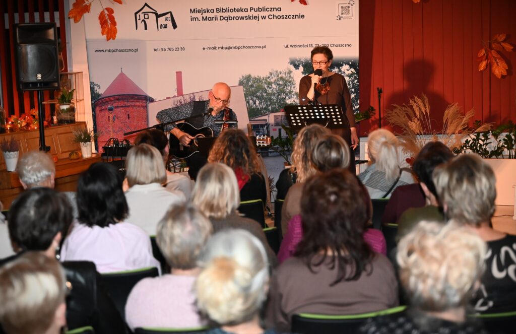 Przed publicznością od lewej: Marek Staniewski, siedzący na krześle i grający na gitarze oraz Marta Staniewska, stojąca przed pulpitem na nuty i śpiewająca do mikrofonu.