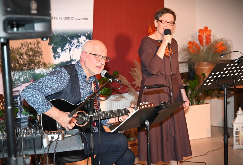 Od lewej: Marek Staniewski, siedzący na krześle i grający na gitarze oraz Marta Staniewska, stojąca przed pulpitem na nuty i śpiewająca do mikrofonu.