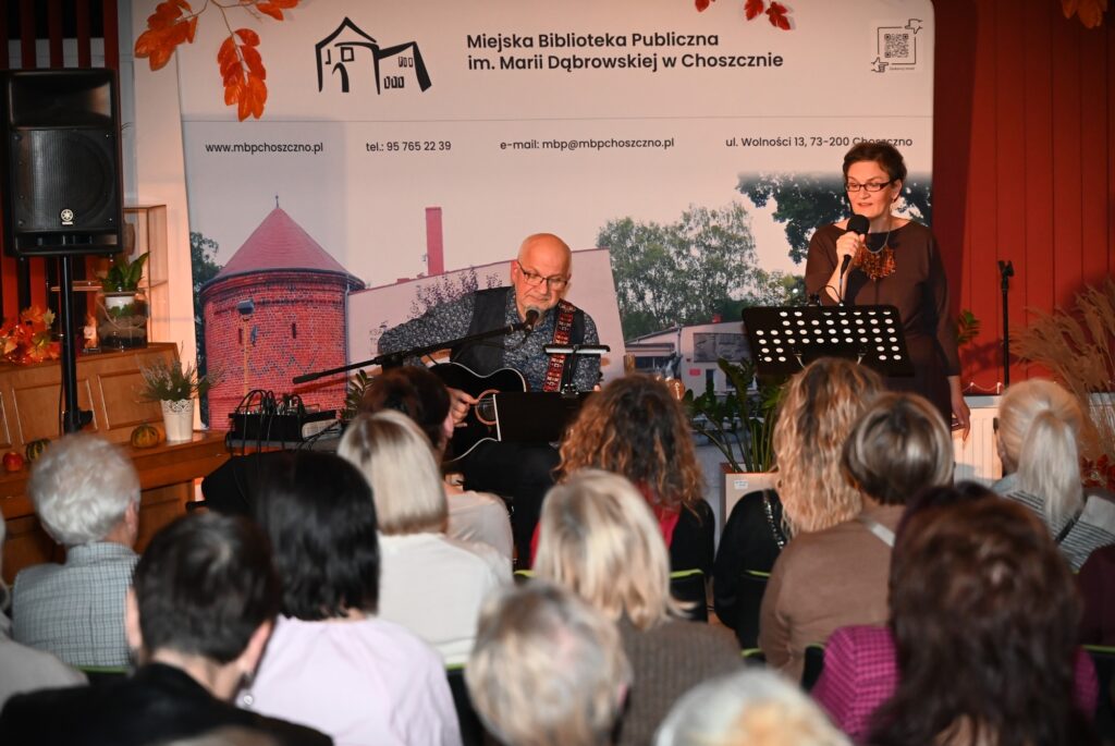 Przed publicznością od lewej: Marek Staniewski, siedzący na krześle i grający na gitarze oraz Marta Staniewska, stojąca przed pulpitem na nuty i śpiewająca do mikrofonu.